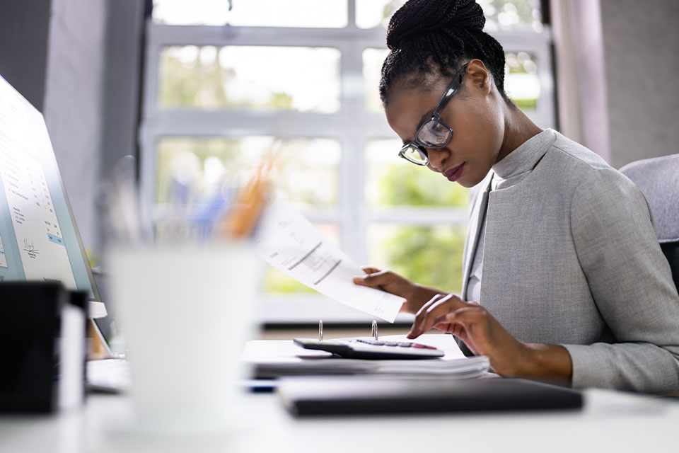 Black woman accountant reviewing paperwork and typing on a calculator in an office setting.