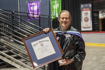 Brad Emmons in commencement regalia, stands with the framed 2026 Crisafullli Award.