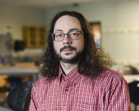 Professor Jonathan Gaffney, with glasses, beard and hair to his shoulders, dressed in a red shirt, stands in his classroom.