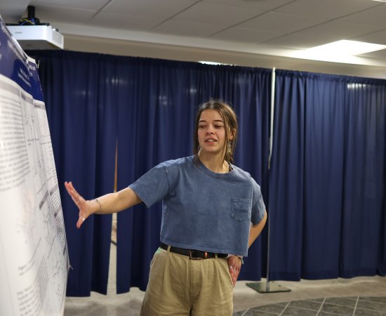 A student makes a presentation beside a poster at the 2024 Student Conference for Research, Professional Activities, and Creative Arts