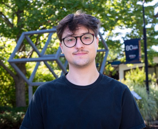 Brayden McCaw in black t-shirt and glasses stands on campus and smiles.