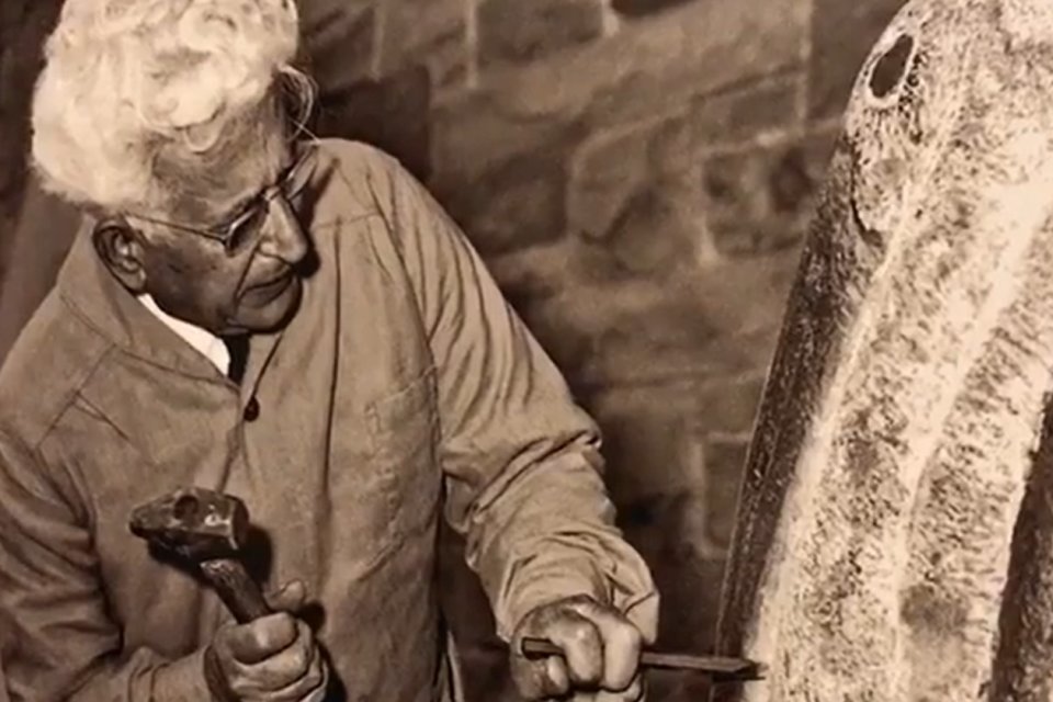 Black and white image of Henry Dispirito at work on a sculpture, with hammer and chisel in hand.