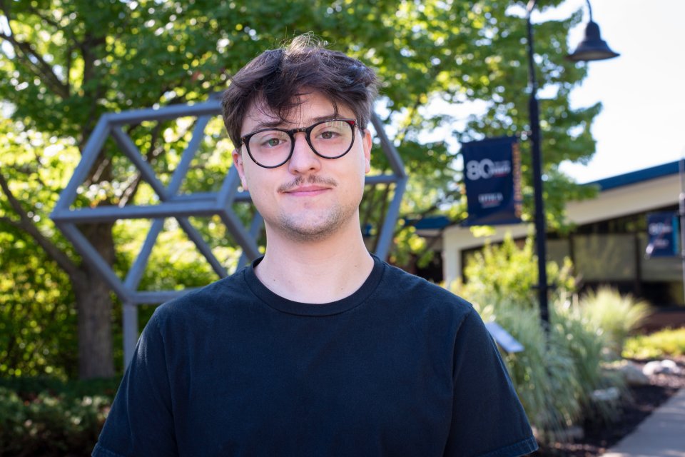 Brayden McCaw in black t-shirt and glasses stands on campus and smiles.